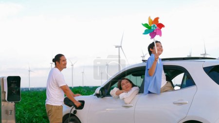 Foto de Concepto de familia feliz progresiva disfrutando de su tiempo en el parque eólico con vehículo eléctrico. Vehículo eléctrico impulsado por energía renovable limpia del generador de aerogeneradores para estación de carga. - Imagen libre de derechos