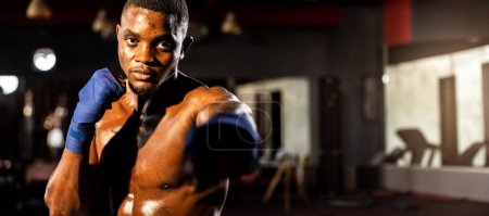 Téléchargez les photos : Combattant de boxe pose torse nu, boxeur africain frapper son poing devant la caméra en position agressive et prêt à se battre à la salle de gym avec sac à pied et équipement de boxe en arrière-plan. Éperon - en image libre de droit