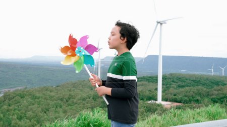 Photo for Progressive young asian boy playing with wind turbine toy in the wind turbine farm, green field over the hill. Green energy from renewable electric wind generator. - Royalty Free Image