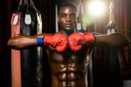 Photo for Boxing fighter posing, African American Black boxer put his hand or fist wearing glove together in front in aggressive stance and ready to fight at gym with kicking bag and boxing equipment. Impetus - Royalty Free Image