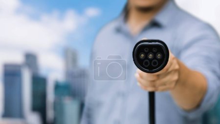 Photo for Focus EV charger pointing in front of camera with businessmans hand in blurry background of cityscape and cloud. Electric car charger plug and alternative clean energy reducing CO2 emission.Peruse - Royalty Free Image