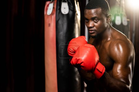 Photo for Boxing fighter shirtless posing, African American Black boxer wearing red glove in defensive guard stance ready to fight and punch at gym with kick bag and boxing equipment in background. Impetus - Royalty Free Image