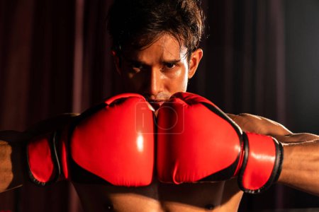 Photo for Muay Thai boxer punch fish his hand together in front of camera in ready to fight stance posing at gym with boxing equipment in background. Focused determination eye and prepare for challenge. Impetus - Royalty Free Image