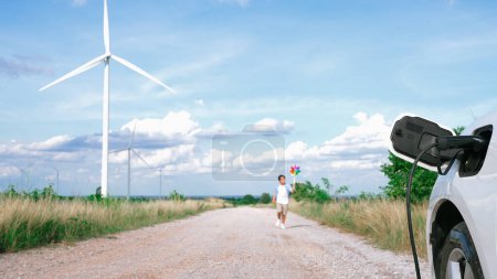 Foto de Niño progresivo jugando juguete molino de viento junto al coche EV en el parque eólico. Generador eléctrico de viento por generador de turbina eólica en el lado del campo. - Imagen libre de derechos