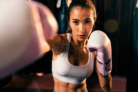 Téléchargez les photos : Femme asiatique Muay boxer thaïlandais poing poing devant la caméra en position de combat prêt à poser à la salle de gym avec équipement de boxe en arrière-plan. Regard de détermination concentré et préparation au défi. Impulsion - en image libre de droit