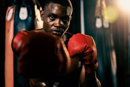 Téléchargez les photos : Combattant de boxe pose torse nu, boxeur noir afro-américain frapper son poing en position agressive et prêt à se battre à la salle de gym avec sac de coups de pied et équipement de boxe en arrière-plan. Impulsion - en image libre de droit