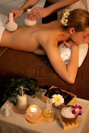 Téléchargez les photos : Chaud à base de plantes balle spa massage du corps, masseur compresse doucement sac à herbes sur le corps de la femme. Tranquille et sérénité des loisirs aromathérapie dans l'éclairage chaud des bougies au salon de spa. Tranquillité - en image libre de droit