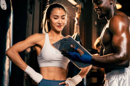 Téléchargez les photos : Femme asiatique Muay boxeuse thaïlandaise et son entraîneur de boxe personnelle discuter de ses progrès physiques dans la salle de gym reflétant son engagement à la croissance musculaire de son corps et la performance de boxe. Impulsion - en image libre de droit