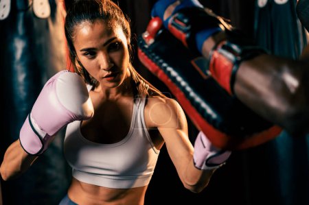 Photo for Asian female Muay Thai boxer punching in fierce boxing training session, delivering strike to her sparring trainer wearing punching mitts, showcasing Muay Thai boxing technique and skill. Impetus - Royalty Free Image