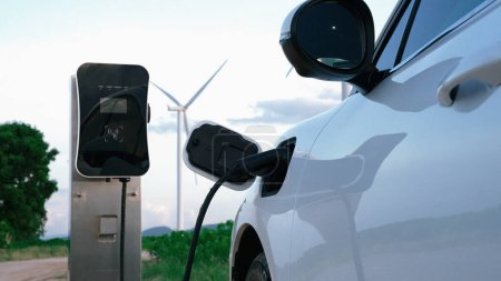 Photo for Progressive future energy infrastructure concept of electric vehicle being charged at charging station powered by green and renewable energy from a wind turbine in order to preserve the environment. - Royalty Free Image