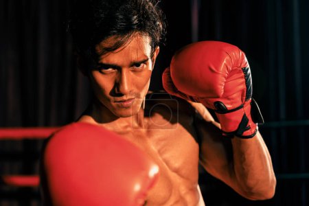 Foto de El boxeador Muay Thai golpea su puño frente a la cámara en posición lista para luchar posando en el gimnasio con equipo de boxeo en segundo plano. Ojos de determinación enfocados y prepararse para el desafío. Impulso - Imagen libre de derechos