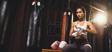 Téléchargez les photos : Femme asiatique déterminée Muay boxeuse thaïlandaise avec muscularité corps de préparation physique enveloppe sa main et dons ou porter un gant de boxe, se préparant à un entraînement intense de boxe sur le ring à la salle de gym. Éperon - en image libre de droit