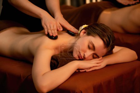 Photo for Hot stone massage at spa salon in luxury resort with warm candle light, blissful couple customer enjoying spa basalt stone massage glide over body with soothing warmth. Quiescent - Royalty Free Image