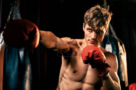 Téléchargez les photos : Combattant de boxe pose torse nu, boxeur caucasien frapper son poing devant la caméra en position agressive et prêt à se battre à la salle de gym avec sac à pied et équipement de boxe en arrière-plan. Impulsion - en image libre de droit