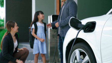 Photo for Progressive parents with electric vehicle and home charging station. Happy family with daughter giving each other high fives before leave for school. Alternative future transportation concept of EVs - Royalty Free Image