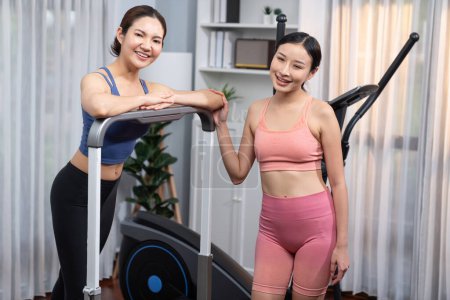 Foto de Mujer asiática atlética enérgica y fuerte corriendo en máquina elíptica en casa con compañero de entrenamiento o entrenador. Búsqueda de físico en forma y compromiso con un estilo de vida saludable. Vigoroso - Imagen libre de derechos