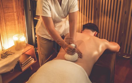 Photo for Hot herbal ball spa massage body treatment, masseur gently compresses herb bag on man body. Tranquil and serenity of aromatherapy recreation in warm lighting of candles at spa salon. Quiescent - Royalty Free Image