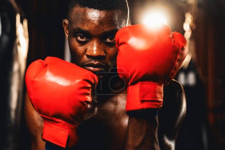 Téléchargez les photos : Combattant de boxe pose torse nu, boxeur noir afro-américain portant un gant rouge en position de garde défensive prêt à se battre et frapper à la salle de gym avec sac de coup de pied et équipement de boxe en arrière-plan. Impulsion - en image libre de droit