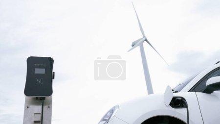 Photo for Progressive future energy infrastructure concept of electric vehicle being charged at charging station powered by green and renewable energy from a wind turbine in order to preserve the environment. - Royalty Free Image