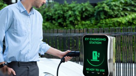 Photo for Businessman pull and hold EV charger plug form electric car charging station at city car park area background. Futuristic clean sustainable energy and EV car technological advancement.Peruse - Royalty Free Image