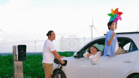 Téléchargez les photos : Concept de famille heureuse progressive appréciant leur temps au parc éolien avec véhicule électrique. Véhicule électrique alimenté par de l'énergie renouvelable propre provenant d'éoliennes pour la station de recharge. - en image libre de droit