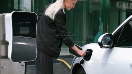 Photo for Progressive businesswoman unplug charger plug from charging station to his electric car before driving around city center. Eco friendly rechargeable car powered by sustainable and clean energy. - Royalty Free Image
