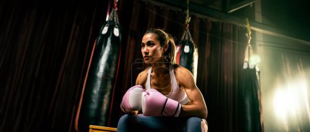 Foto de Mujer asiática Muay Thai boxeador o kickboxing tomando un breve descanso sentado con sus guantes en el gimnasio con equipo de boxeo en el fondo. Deportiva corporal fuerte y musculosa. Espuela - Imagen libre de derechos