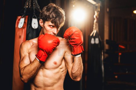 Téléchargez les photos : Combattant de boxe pose torse nu, boxeur homme caucasien portant un gant rouge en position de garde défensive prêt à se battre et frapper à la salle de gym avec sac de coup de pied et équipement de boxe en arrière-plan. Impulsion - en image libre de droit