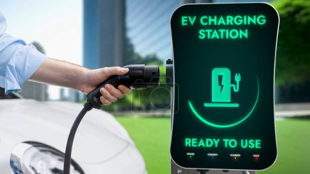 Photo for Businessman pull EV charger to recharge his electric cars battery from charging station in green eco city park background. Future innovative EV car and energy sustainability. Peruse - Royalty Free Image