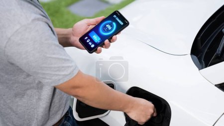 Foto de Inserte el cargador de EV a mano en el coche eléctrico para recargar la batería mientras comprueba la visualización del estado de la batería en el teléfono inteligente. Futuro energía sostenible y limpia para el coche EV. Período - Imagen libre de derechos