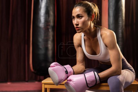 Téléchargez les photos : Femme asiatique Muay boxeuse thaïlandaise ou kickboxing prendre une courte pause assis avec ses gants sur la salle de gym avec du matériel de boxe en arrière-plan. Forte et musclée sportive du corps. Impulsion - en image libre de droit