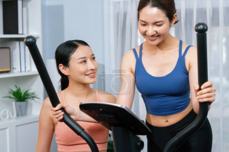 Téléchargez les photos : Femme asiatique athlétique énergique et forte fonctionnant sur une machine elliptique à la maison avec un copain ou un entraîneur d'entraînement. Poursuite d'ajustement physique et l'engagement à un mode de vie sain. Vigoureux - en image libre de droit