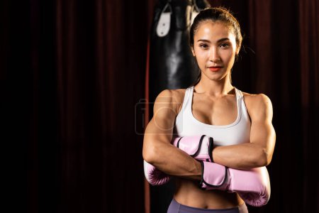 Photo for Asian female Muay Thai boxer punch fist in front of camera in ready to fight stance posing at gym with boxing equipment in background. Focused determination eyes and prepare for challenge. Impetus - Royalty Free Image