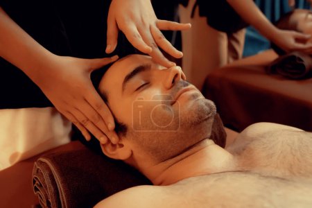 Téléchargez les photos : Homme caucasien profiter relaxant anti-stress massage de la tête et choyer beauté faciale loisirs de la peau dans la bougie chaude éclairage spa salon ambiant dans un complexe de luxe ou un hôtel. Tranquillité - en image libre de droit