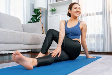 Foto de Mujer asiática en ropa deportiva haciendo ejercicio de yoga en la alfombra de fitness como su rutina de entrenamiento en casa. Cuidado corporal saludable y meditación tranquila en el estilo de vida del yoga con comodidad y relajación. Vigoroso - Imagen libre de derechos