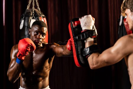 Téléchargez les photos : Boxer noir afro-américain frappant à l'entraîneur ou entraîneur portant des mitaines de poinçonnage comme équipement d'entraînement de sac de boxe dans la salle de gym. Entraînement de force et d'endurance pour un match de boxe professionnel. Impulsion - en image libre de droit