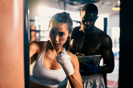 Photo for Asian female muay thai boxer with muscular and strong physical body training, punching at boxing bag instructed by her personal trainer or coach in the gym. Martial art and sport concept. Impetus - Royalty Free Image