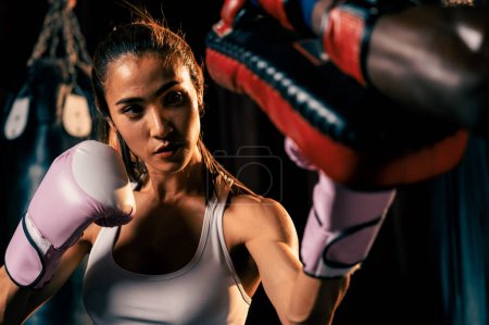 Téléchargez les photos : Femme asiatique Muay boxer thaïlandais poinçonnage dans la séance d'entraînement de boxe féroce, la livraison de grève à son entraîneur sparring portant des mitaines de poinçonnage, mettant en valeur la technique et l'habileté Muay boxe thaïlandaise. Impulsion - en image libre de droit