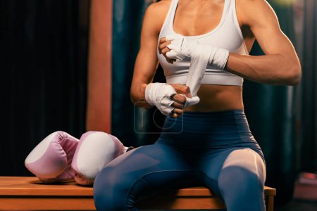 Foto de La decidida boxeadora asiática Muay Thai con cuerpo de musculatura y preparación física envuelve su mano y se pone o usa guante de boxeo, preparándose para un intenso entrenamiento de boxeo en el ring en el gimnasio. Impulso - Imagen libre de derechos