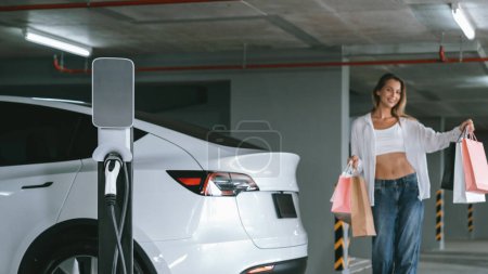 Foto de Mujer joven viaja con coche eléctrico EV al estacionamiento del centro comercial carga en el centro de la ciudad mostrando estilo de vida de sostenibilidad urbana por energía verde recargable limpia de entradas de vehículos eléctricos - Imagen libre de derechos