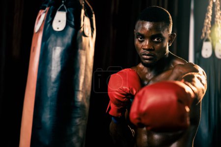 Foto de Combatiente de boxeo posando sin camisa, boxeador negro afroamericano golpea su puño en postura agresiva y listo para luchar en el gimnasio con bolsa de patadas y equipo de boxeo en segundo plano. Impulso - Imagen libre de derechos