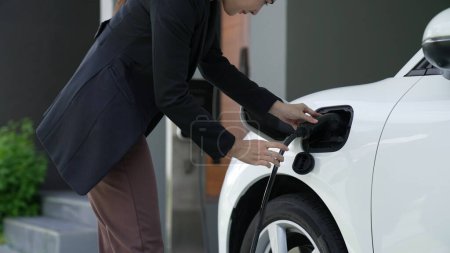 Foto de Mujer progresista instala un enchufe de la estación de carga en su vehículo eléctrico en casa. Automóviles EV proporcionan un concepto ambientalmente beneficioso de energía limpia y verde. - Imagen libre de derechos