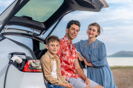 Photo for Family vacation trip traveling by the beach with electric car, lovely family sit on the trunk, charging EV car battery with green and sustainable energy. Family travel and eco-friendly car. Perpetual - Royalty Free Image
