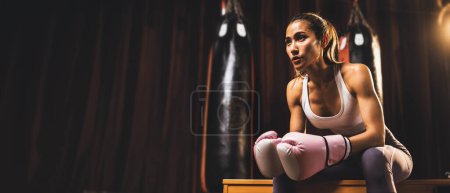 Photo for Asian female Muay Thai boxer or kickboxing taking short break sitting with her gloves on at the gym with boxing equipment in background. Strong and muscular body sportswoman. Spur - Royalty Free Image