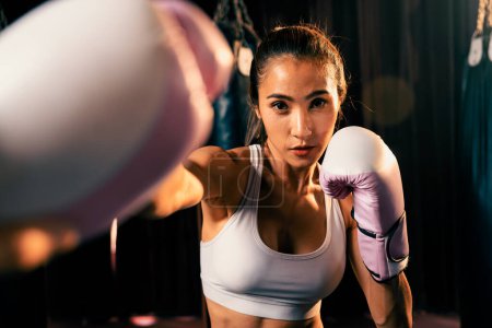 Téléchargez les photos : Femme asiatique Muay boxer thaïlandais poing poing devant la caméra en position de combat prêt à poser à la salle de gym avec équipement de boxe en arrière-plan. Regard de détermination concentré et préparation au défi. Impulsion - en image libre de droit