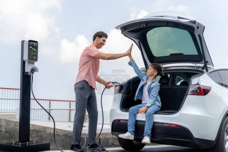 Photo for Family road trip vacation traveling by the sea with electric car, father and son high five after reach destination at EV charging station by the seashore. Eco-friendly car for environment. Perpetual - Royalty Free Image