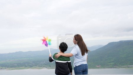 Foto de Una mujer progresista y su hijo están de vacaciones, disfrutando de la belleza natural de un lago en el fondo de una colina mientras el niño lleva un molino de viento de juguete. - Imagen libre de derechos