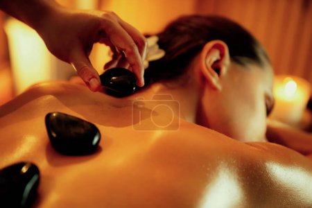 Photo for Closeup hot stone massage at spa salon in luxury resort with warm candle light, blissful woman customer enjoying spa basalt stone massage glide over body with soothing warmth. Quiescent - Royalty Free Image