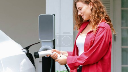 Téléchargez les photos : Femme écologique moderne rechargeant le véhicule électrique de la station de charge EV à la maison. Technologie de voiture EV utilisé pour les résidents de la maison à la durabilité environnementale future. Synchronos - en image libre de droit