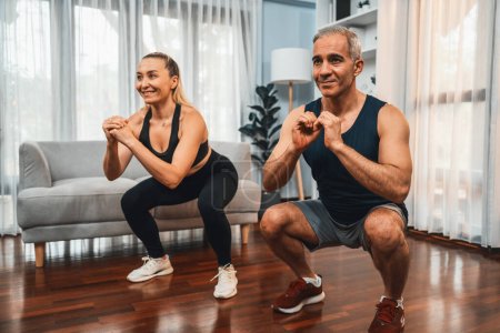 Téléchargez les photos : Couple de personnes âgées athlétiques et sportives s'engageant dans une séance d'entraînement de jour de jambe avec squat ensemble à la maison l'exercice comme concept de mode de vie sain de corps de forme après la retraite. Clout - en image libre de droit